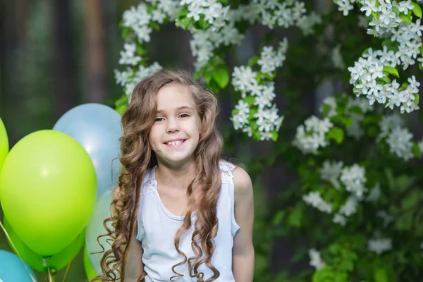 Маленькая красивая улыбающаяся девочка с длинными вьющимися волосами, одетая в умную пушистую юбку, играющую в весеннем саду, держа в руках большую связку шариков. Концепция счастливого детства — стоковое фото