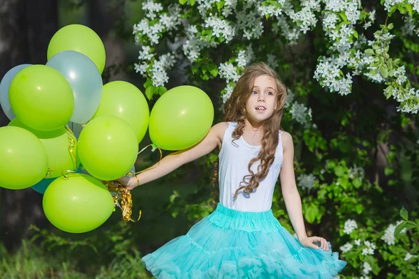 Pequeña chica sonriente hermosa con el pelo rizado largo usando falda mullida inteligente jugando en el jardín de primavera sosteniendo en las manos un gran montón de globos. Concepto de infancia feliz — Foto de Stock