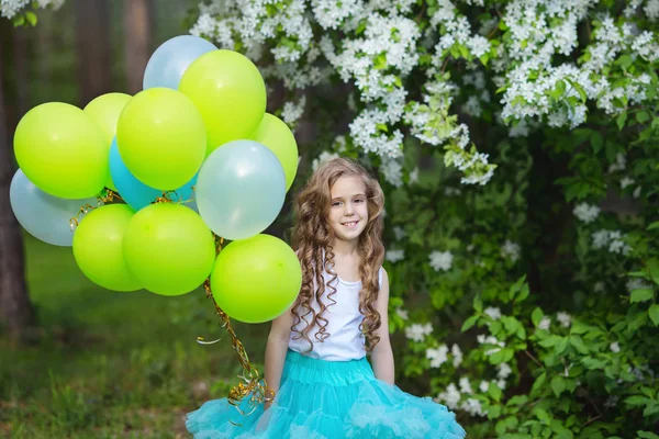 Маленькая красивая улыбающаяся девочка с длинными вьющимися волосами, одетая в умную пушистую юбку, играющую в весеннем саду, держа в руках большую связку шариков. Концепция счастливого детства — стоковое фото