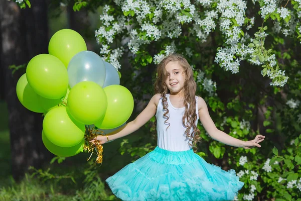 Pequena menina sorridente bonita com cabelos longos encaracolados vestindo saia fofa inteligente jogando no jardim da primavera segurando nas mãos um grande monte de balões. Conceito de infância feliz — Fotografia de Stock
