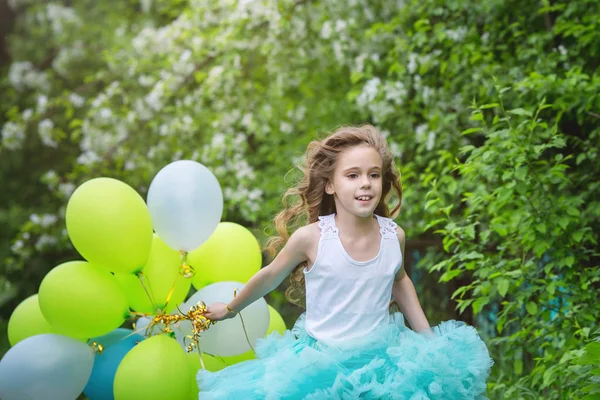 Pequena menina sorridente bonita com cabelos longos encaracolados vestindo saia fofa inteligente jogando no jardim da primavera segurando nas mãos um grande monte de balões. Conceito de infância feliz — Fotografia de Stock