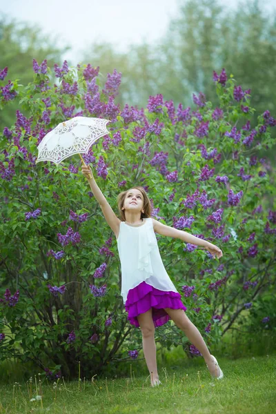 Красивая маленькая девочка в белом платье с кружевным зонтиком играет с кружевным зонтиком на фоне весеннего сиреневого сада — стоковое фото