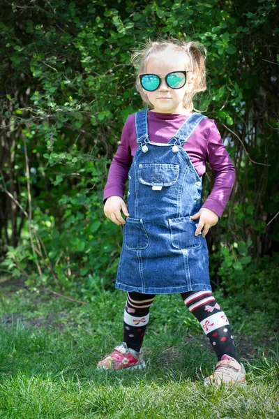 Uma criança de óculos e um sarafan do caton. Um modelo pequeno. Na moda. Menina posando ao ar livre — Fotografia de Stock