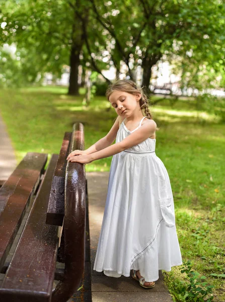 A beautiful little girl in a white long dress staying near the bench in the park with her eyes closed — Stock Photo, Image