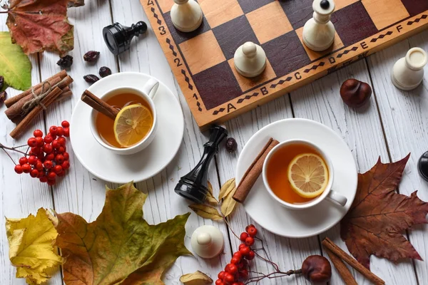 A cozy photo with two cups of tea, board with unfinished chess game, autumn leaves and cinnamon sticks