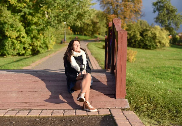 Uma foto de uma jovem garota sorridente sentada em uma ponte de madeira em um parque e desfrutando do sol de outono com os olhos fechados — Fotografia de Stock