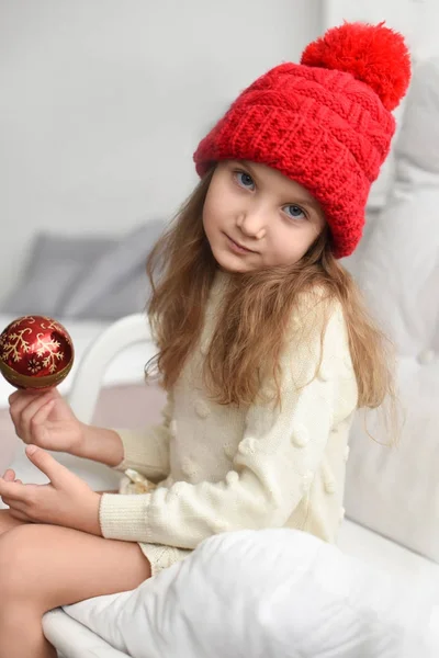 An indoor portrait of a little girl in a knitted warm red cap holding Christmas decorations - a photo representing a concept of waiting for Christmas — Stock Photo, Image