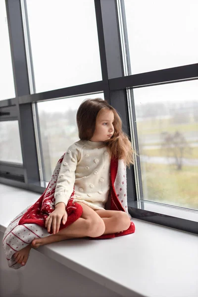 Una niña se envolvió en una acogedora manta de punto y se sentó en una repisa de la ventana mirando a través del vidrio Imágenes de stock libres de derechos