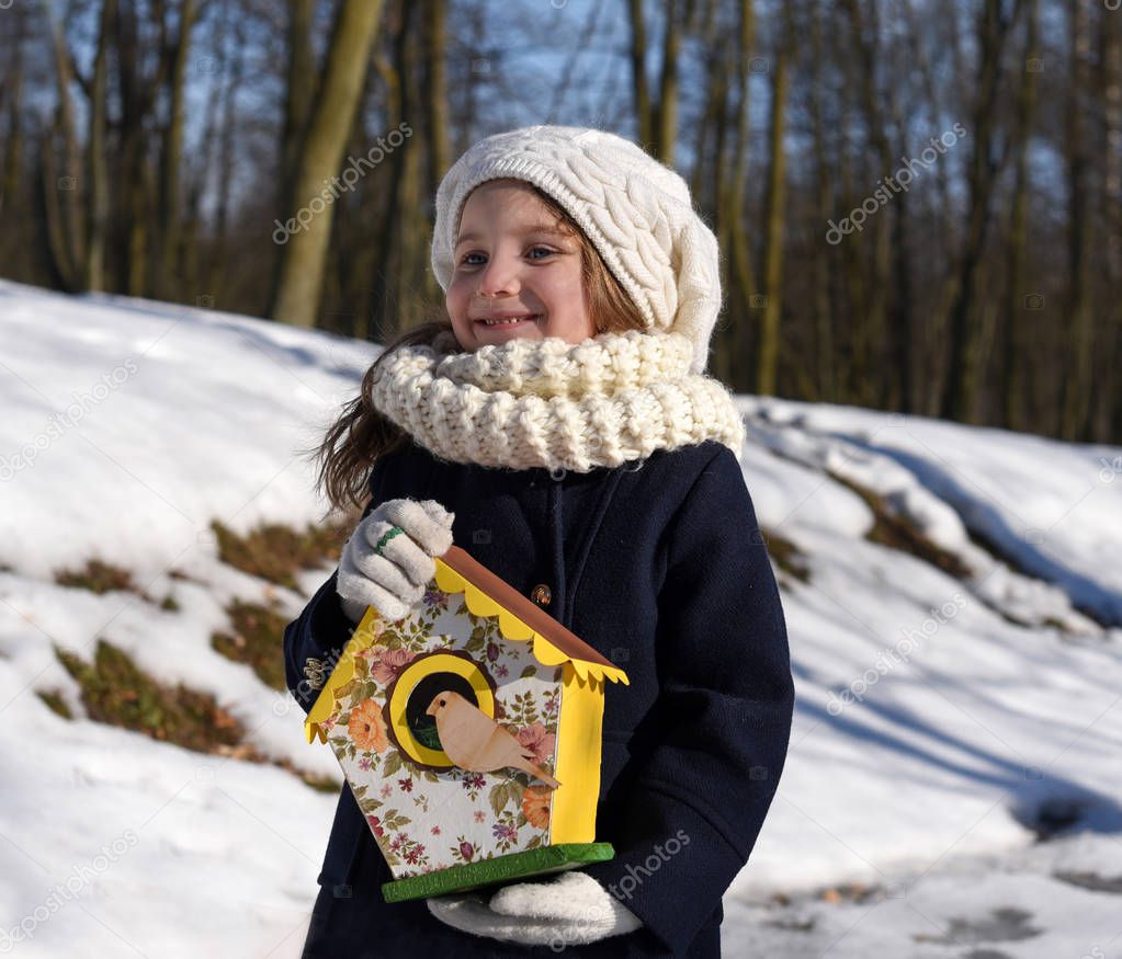 An early spring photo of a beautiful smiling little girl holding a bird house in a park
