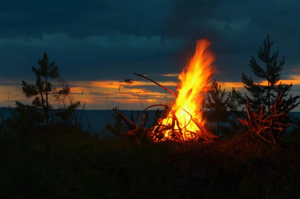 Grand feu de joie nocturne près de la rivière au coucher du soleil . Photo De Stock