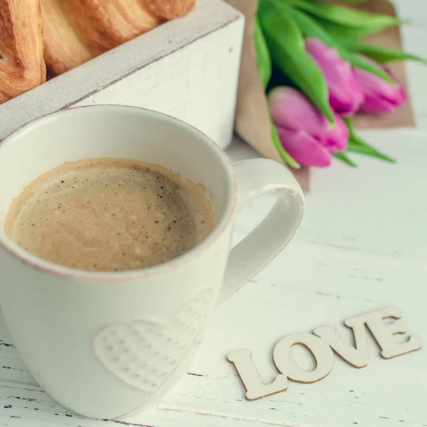 Xícara de café com croissants, buquê de tulipas rosa e palavra de madeira AMOR — Fotografia de Stock