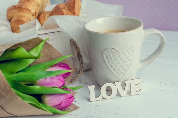 Kopp kaffe och croissanter, bukett av rosa tulpaner och trä ordet kärlek — Stockfoto