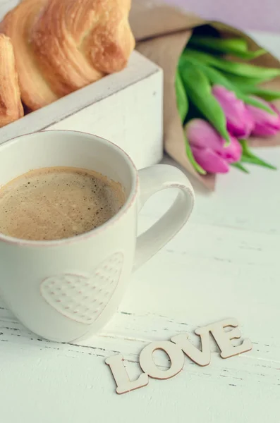 Xícara de café com croissants, buquê de tulipas rosa e palavra de madeira AMOR — Fotografia de Stock
