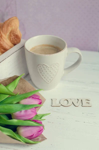 Kopp kaffe och croissanter, bukett av rosa tulpaner och trä ordet kärlek — Stockfoto