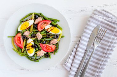 Yaz sıcak salata pişmiş fasulye, ton balığı, domates, haşlanmış yumurta ve sos Balzamico glassa bıçak ve çatal ahşap arka plan üzerinde beyaz plaka ile. Sağlıklı beslenme kavramı. Üstten Görünüm.