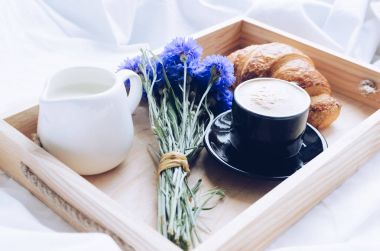 Romantik yaz odaya yatak, taze kruvasan ile tepsi, fincan sütlü kahve espresso ve mavi Peygamber buket. Günaydın kavramı. Yavaş hayattan zevk. Geleneksel kahvaltı.