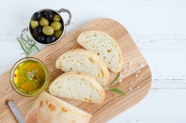 Biberiye, tuz, zeytin ve zeytinyağı ile beyaz rustik tabloda ahşap kesme tahtası üzerinde taze pişmiş ciabatta ekmeği dilimlenmiş. İtalyan gıda kavramı. Üstten Görünüm.