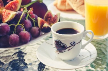 Kahve, bir bardak taze portakal suyu, bir tabak bordo üzüm, incir ve terasta açık havada yaz sabah kruvasan birkaç ile kahvaltı masası. Bir fincan kahve ön planda.