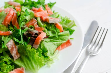 Yaprak sebze salatası salata ile mix, roka ve bıçak ve çatal Beyaz ahşap zemin üzerinde bir plaka üzerinde somon füme. Sağlıklı beslenme kavramı.