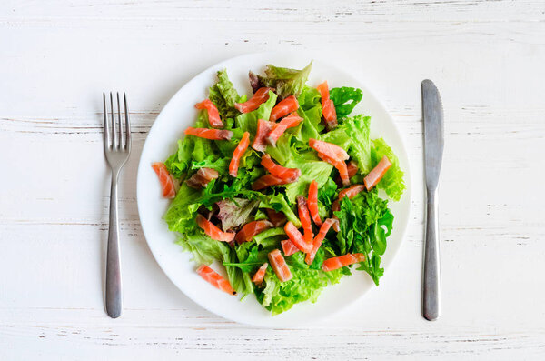 Leaf vegetable salad with salad mix, arugula and smoked salmon on a plate with knife and fork on white wooden background. Healthy food concept. Top view.