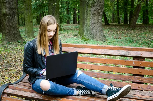 Student studying outdoors. Young woman keyboarding on laptop computer while sitting on the bench in park. Blonde female teen working on net-book after her lectures in University. Copy space for text.