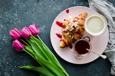 Bir fincan kahve, kruvasan ve siyah arka planda pembe lalelerle reçel. Anneler Günü 'nde, Sevgililer Günü' nde ya da Kadınlar Günü 'nde kahvaltı. Romantik sabah konsepti sürprizi. Üst görünüm.