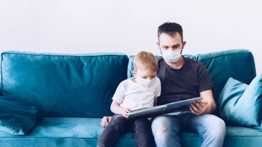 Genç, karantinaya alınmış tıbbi maskeli Coronavirus ailesi kitap okuyor. Corona Virüsü ev karantinası sırasında çocuklarınızla evde etkinlikler. Evde kal. Baba ve oğul milenyumu.