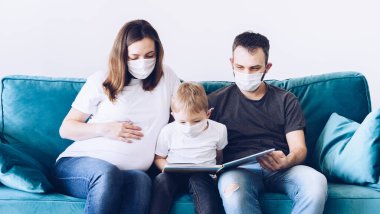 Genç, karantinaya alınmış tıbbi maskeli Coronavirus ailesi kitap okuyor. Corona Virüsü ev karantinası sırasında çocuklarınızla etkinlikler. Evde kal. Baba, hamile anne ve oğul milenyumu..