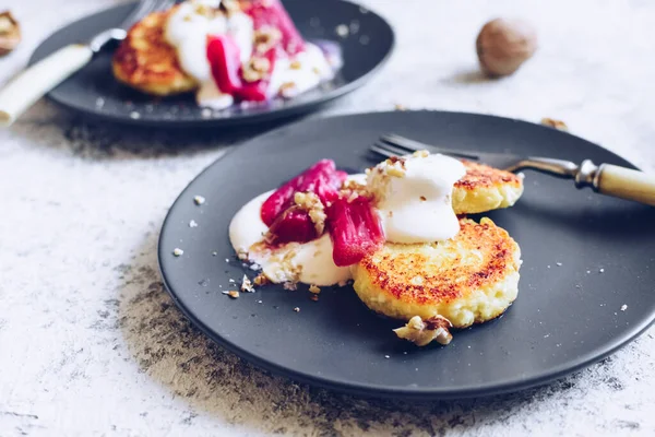 Cottage cheese pancakes served with poached rhubarb on grey background. Gourmet healthy breakfast - cottage cheese syrniki, curd fritters with rhubarb, creme and nuts.