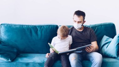 Genç, karantinaya alınmış tıbbi maskeli Coronavirus ailesi kitap okuyor. Corona Virüsü ev karantinası sırasında çocuklarınızla evde etkinlikler. Evde kal. Baba ve oğul milenyumu.