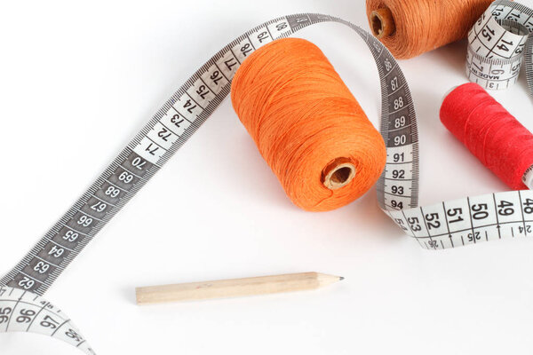 Accessories for sewing: thread, a pencil and a centimeter on a white background. Orange, brown and red bobbin thread.