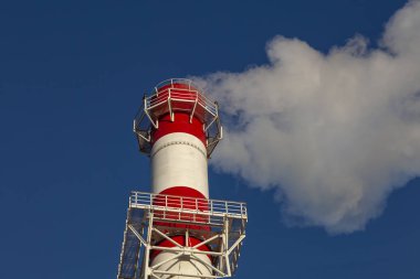 Açık mavi gökyüzüne karşı beyaz ve kırmızı renkli dumanlı fabrika bacası.