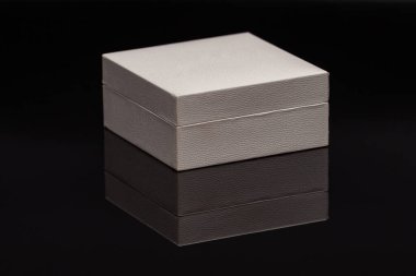 Beyaz kare bir hediye kutusu, kumaşlı bir kaplama siyah bir cam üzerinde duruyor, yansıması görülebiliyor.