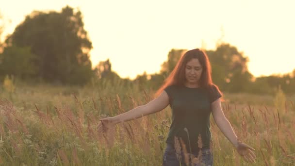 Vacker modell flicka klädd i jeans och en grön skjorta på fältet i solljus. Går till kameran och ler sött. — Stockvideo