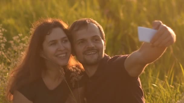 Glückliches romantisches Paar, das bei Sonnenuntergang im Park spaziert und ein Selfie macht. Ein junger Mann küsst seine Freundin nach einem Selfie. — Stockvideo