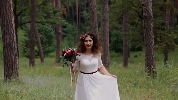 Hermosa novia en un vestido vintage con ramo de colores de flores frescas de cuervo en su cabeza al aire libre en el bosque de verano. Va a la cámara y sonríe . — Vídeos de Stock