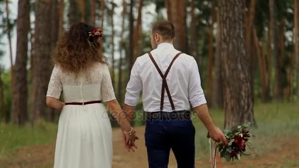 Hermosa pareja ir en un camino forestal. El novio sostiene una mano a la novia, la otra mano un hermoso ramo. La vista desde atrás — Vídeos de Stock