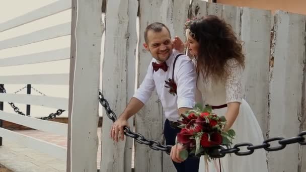 Beautiful pair of hipsters are leaning on the iron chain. Young woman holding a magnificent bouquet. On the background of old wooden fence — Stock Video
