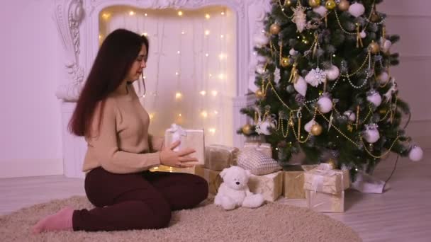 Mooie jonge vrouw in de buurt van open haard in kerst zet een geschenk onder de kerstboom voor haar minnaar. — Stockvideo