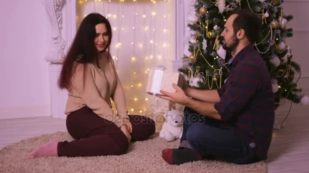 Un joven regala a su novia para Navidad. Los amantes se sientan frente a la chimenea en la alfombra beige . — Vídeo de stock