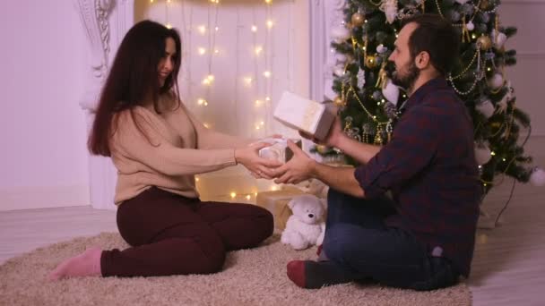 Jovem casal feliz sentado ao lado da lareira dando presentes uns aos outros. Feliz Natal! . — Vídeo de Stock