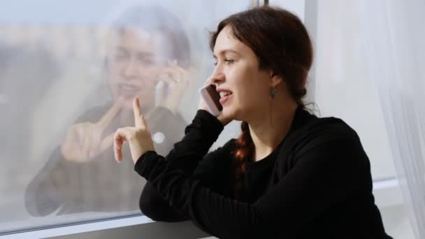 一个年轻女子在电话上愉快地交谈，手指在窗户上画. — 图库视频影像