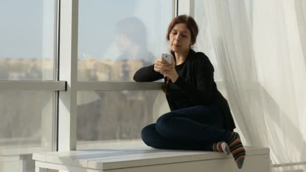 Glückliche junge Frau schreibt SMS in sozialen Netzwerken, während sie am Fenster sitzt — Stockvideo
