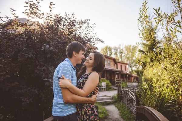 Pasangan romantis yang bahagia di desa, berjalan-jalan di jembatan kayu. Wanita cantik muda dalam gaun musim panas panjang memeluk pria . — Stok Foto