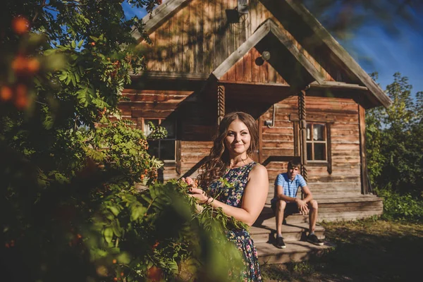 Loving romantic couple in the village, near a wooden house. A man sits on the porch, a young woman in the foreground. Concept: love, romance, summer. — Stock Photo, Image