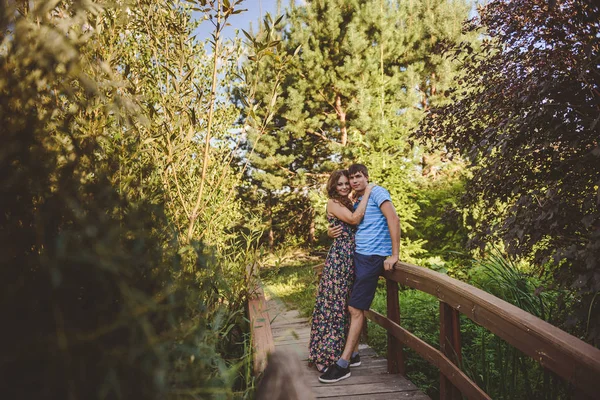 Pasangan romantis yang bahagia di desa, berjalan-jalan di jembatan kayu. Wanita cantik muda dengan gaun musim panas yang panjang memeluk pria itu. Lihat ke kamera. . — Stok Foto