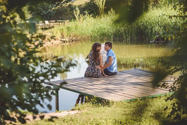 Gelukkige romantisch paar op een houten brug in de buurt van het meer. Man en een jonge vrouw praten zittend op de oever van het meer. — Stockfoto