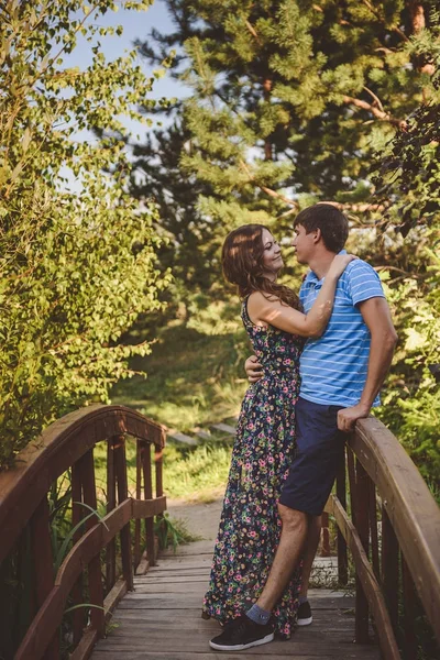 Feliz pareja romántica en el pueblo, pasear por el puente de madera. Joven hermosa mujer en vestido de verano largo abrazando al hombre . — Foto de Stock