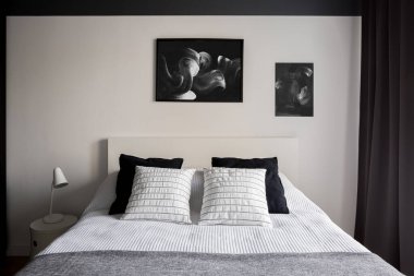 Siyah beyaz yatak odası, duvarında sanat eserleri ve komodinin üzerinde basit beyaz lamba.