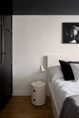 Siyah ve beyaz yatak odasının içinde büyük siyah gardırop ve beyaz komodinin üzerinde basit beyaz lamba var.
